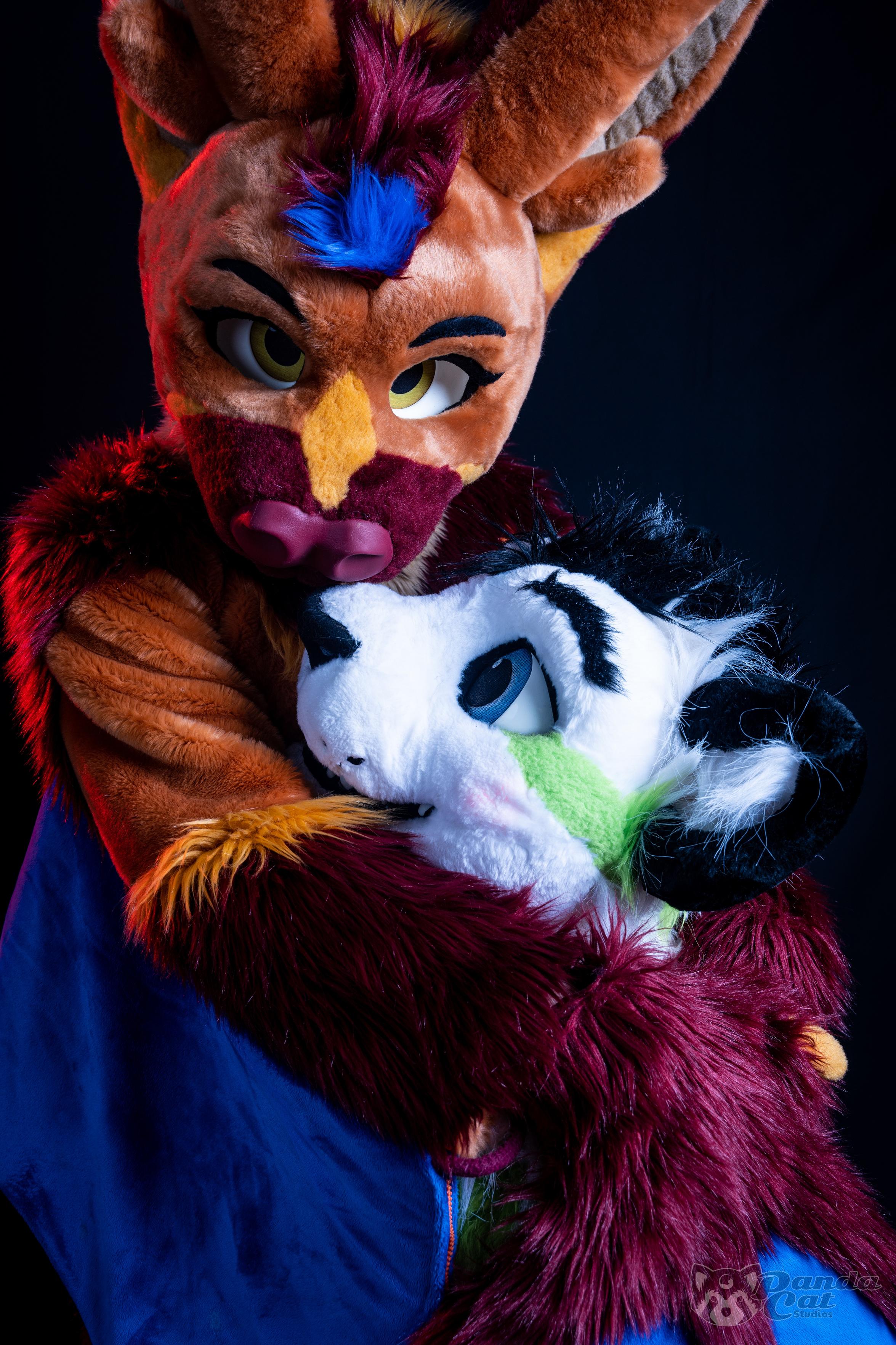 The picture is dominated by a brown bat with dark-red accents on their muzzle, ears and forearms. They are looking right at the camera with a sly attitude. Their arms, with dark-blue wings, are wrapped around a white tiger with green stripes, only his head visible from over the bat's arms, looking up at them while blushing slightly.